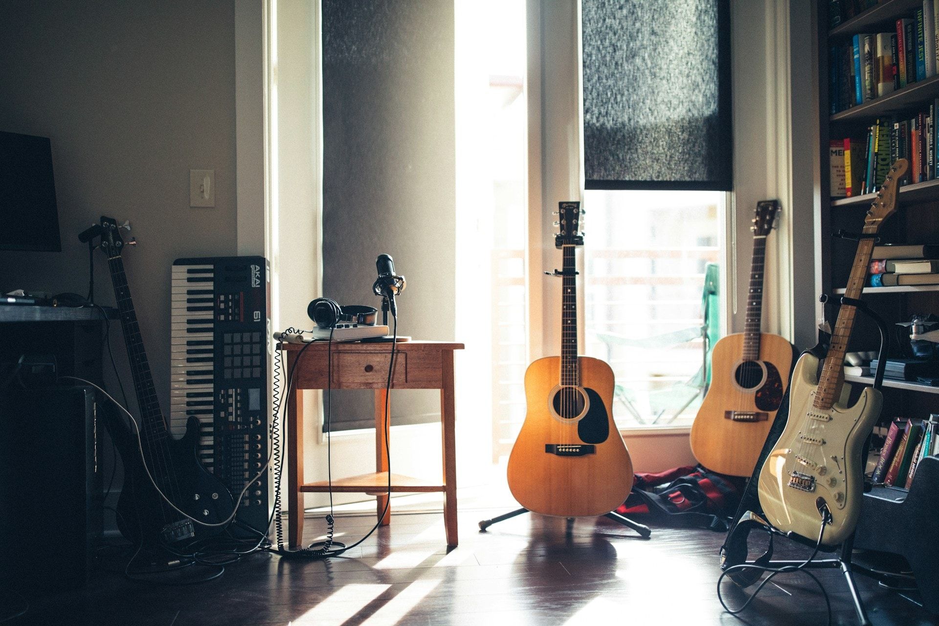 Sala clara com três violões e uma guitarra em suportes, um teclado e uma mesinha de madeira com fone de ouvido, ao lado de uma janela.